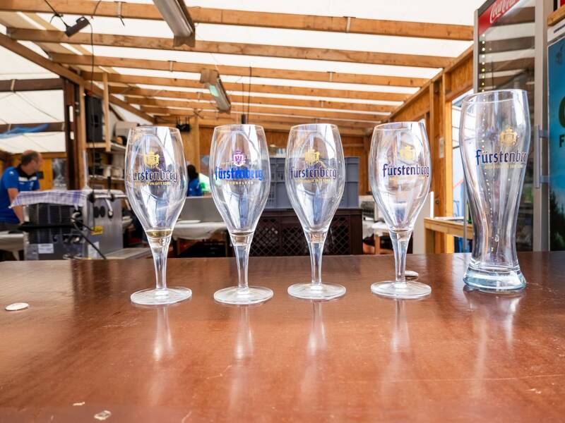 Fünf leere Fürstenberg-Biergläser stehen auf einer Theke in einem überdachten, hölzernen Verkaufsstand.