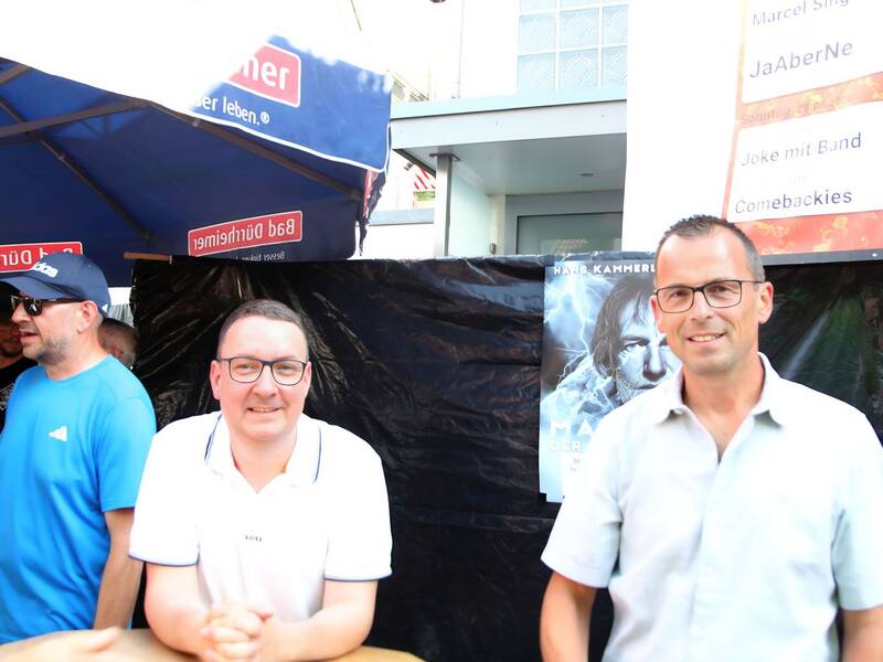 Zwei Männer in Freizeitkleidung stehen unter einem blauen Schirm vor einem schwarzen Hintergrund mit Postern.