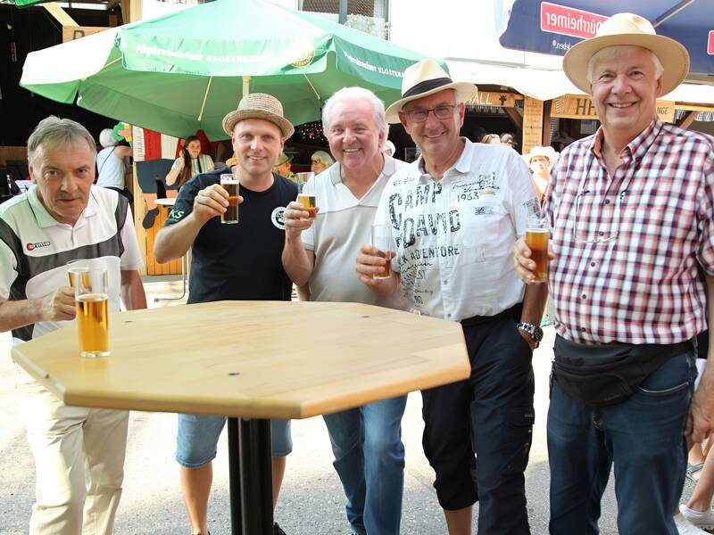 Fünf Männer stehen um einen Stehtisch im Freien, halten Biergläser und lächeln in die Kamera, während im Hintergrund Menschen und Sonnenschirme zu sehen sind.