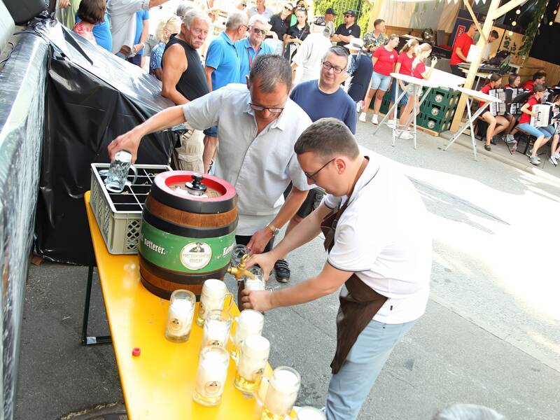 Ein Mann zapft Bier aus einem Fass in Gläser bei einem Straßenfest, umgeben von Menschen und einer Blasmusikgruppe im Hintergrund.