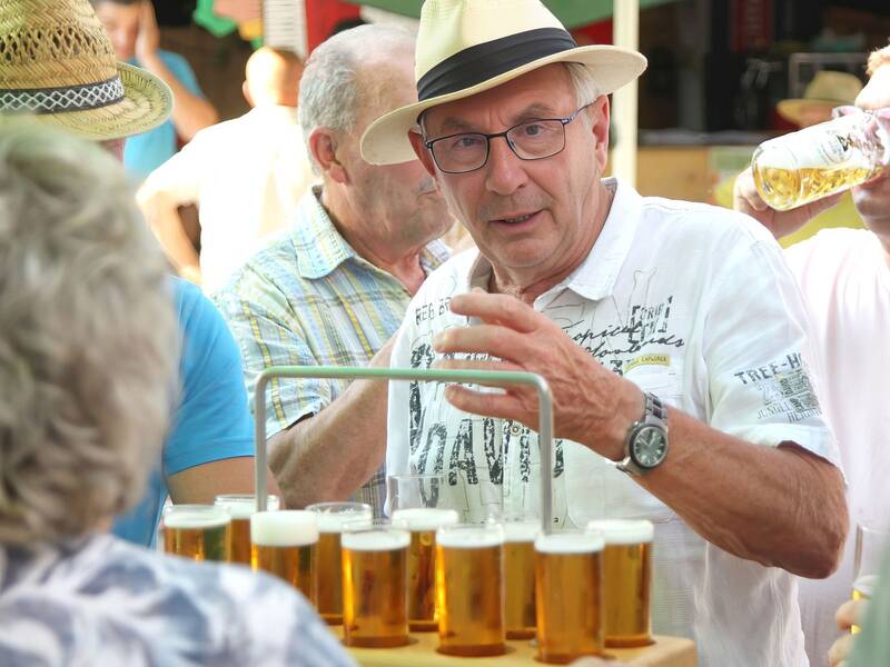 Eine Gruppe von Männern steht bei einer Veranstaltung im Freien, wobei ein Mann mit Hut und weißem Hemd einen Getränkehalter mit mehreren Biergläsern hält.
