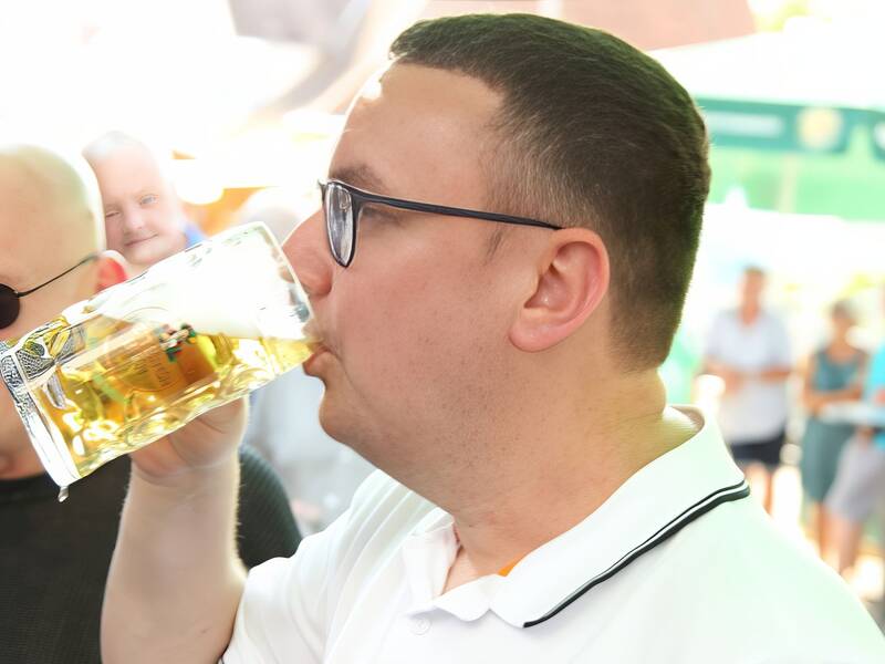 Ein Mann mit Brille trinkt ein großes Glas Bier, während er in einer geselligen Umgebung steht.