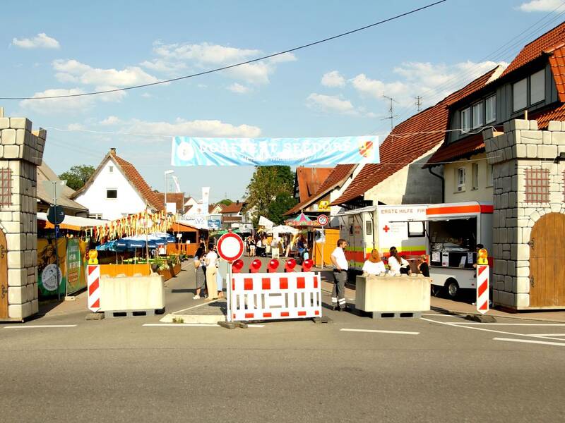 Der Eingang zu einem Fest wird durch eine Barriere mit Burgmauern und einem Banner darüber markiert, während im Hintergrund Menschen und Verkaufsstände zu sehen sind.