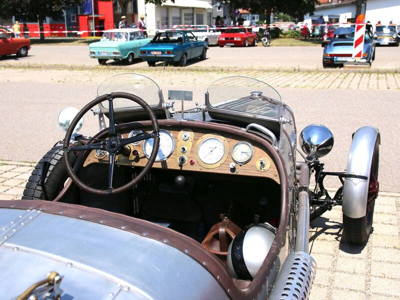 Das Bild zeigt den Innenraum eines klassischen Oldtimers mit sichtbarem Holzarmaturenbrett, Lenkrad und Rückspiegel, umgeben von mehreren anderen Oldtimern auf einem Parkplatz.