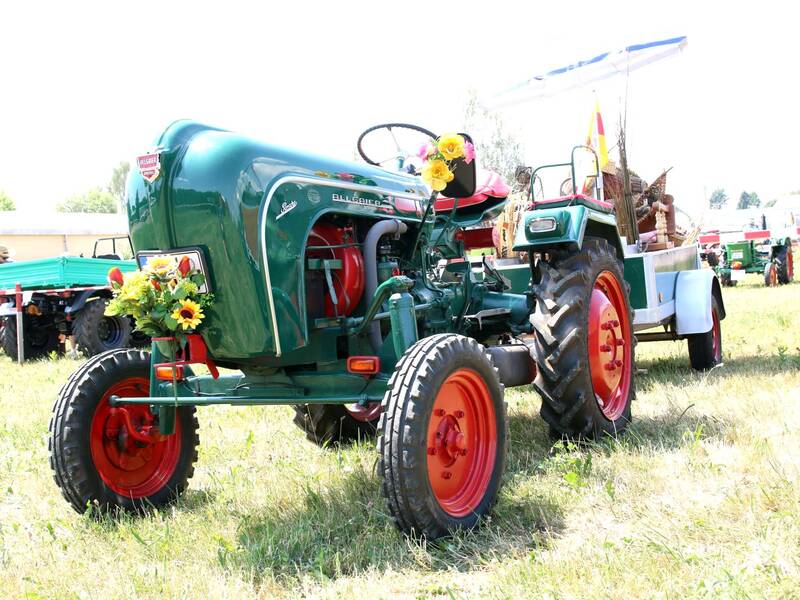 Ein grüner, geschmückter Traktor mit roten Rädern zieht einen Anhänger über eine Wiese.