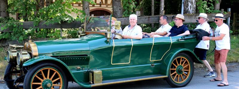 Personen sitzen in einem grünen Oldtimer, während eine Person das Auto von hinten schiebt, vor einer Kulisse aus Bäumen und einem Fachwerkhaus.