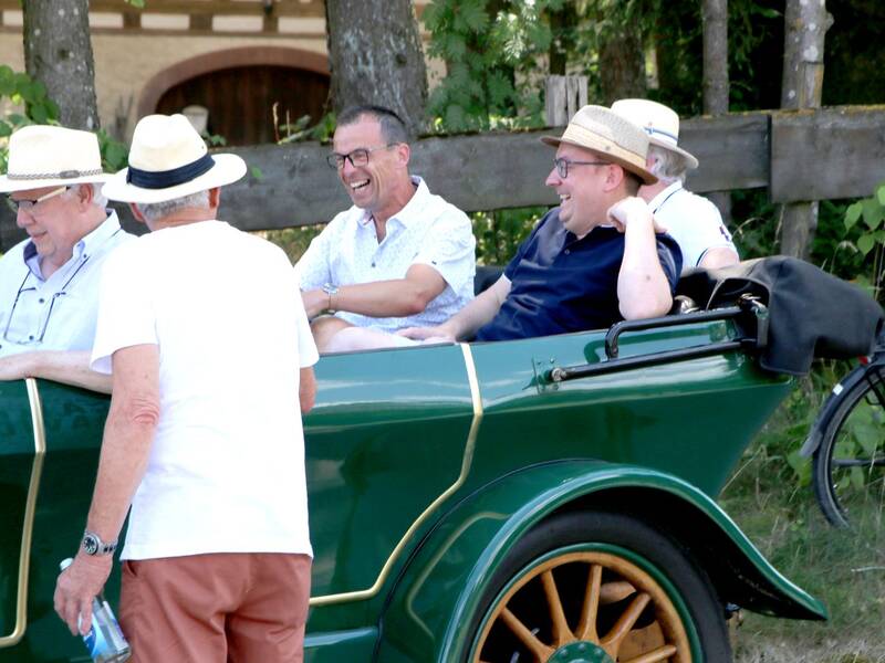 Eine Gruppe von Männern sitzt lachend in einem grünen Oldtimer, einige tragen Strohhüte.