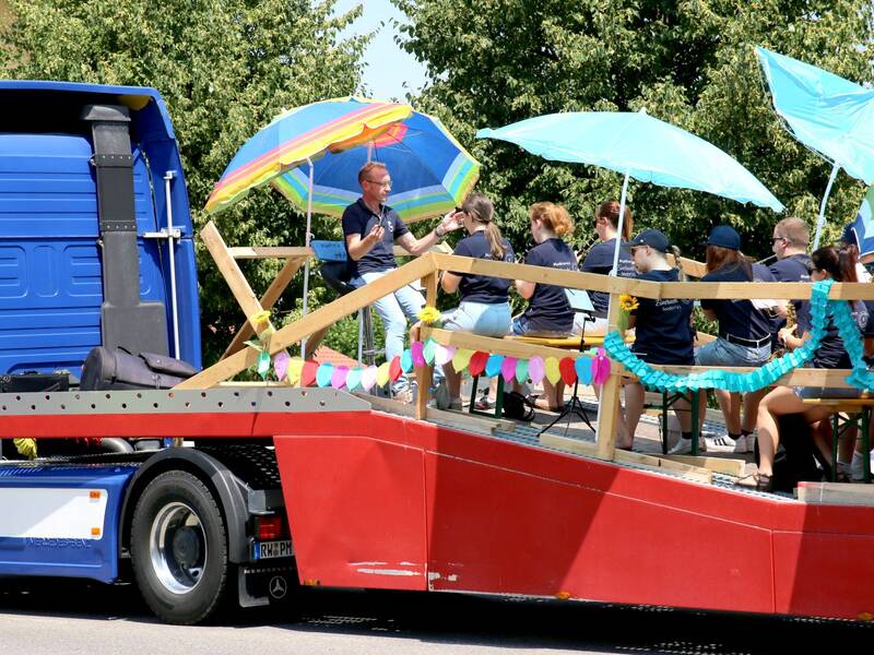 Eine Gruppe von Menschen sitzt unter bunten Sonnenschirmen auf einem dekorierten Anhänger, der von einem blauen LKW gezogen wird.