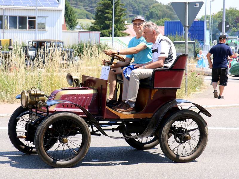 Zwei Männer fahren in einem alten, offenen Automobil über eine Straße.