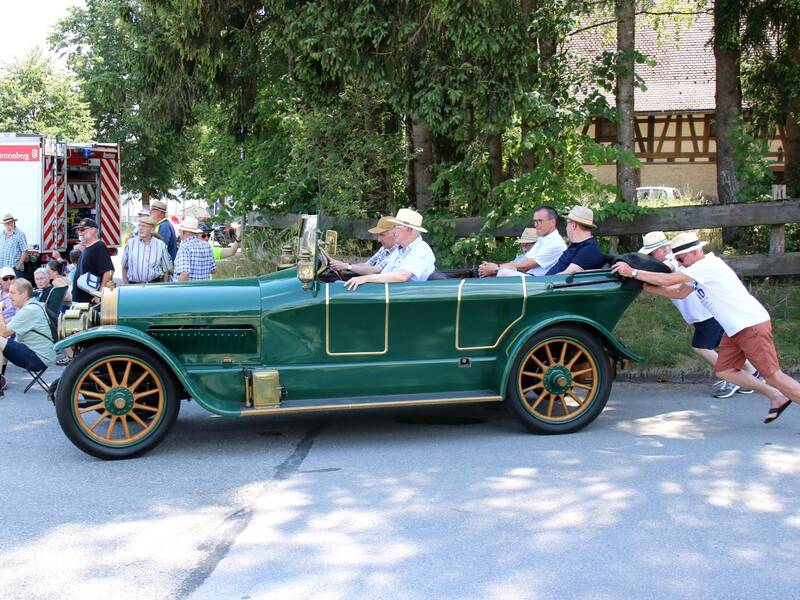 Eine Gruppe von Menschen in einem offenen, grünen Oldtimer-Auto wird von einer weiteren Person angeschoben, während ein Einsatzfahrzeug und Zuschauer im Hintergrund zu sehen sind.