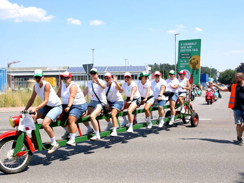 Eine Gruppe von Menschen sitzt auf einem langen, mehrsitzigen Fahrrad in Rot und Grün und fährt auf einer Straße, während eine Person in einer orangefarbenen Weste daneben geht.