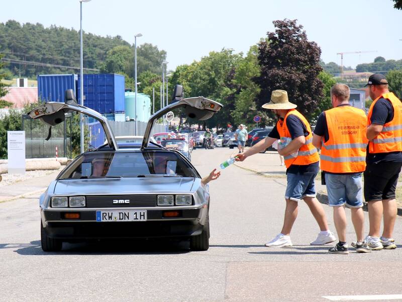 Ein silberner DeLorean mit geöffneten Flügeltüren hält auf einer Straße, während eine Person in einer orangefarbenen Warnweste eine Wasserflasche durch das Fenster reicht.