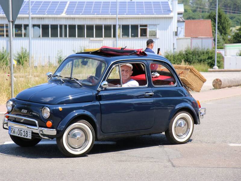 Ein lächelnder Mann fährt in einem blauen Oldtimer mit geöffnetem Verdeck und einem Korb auf der Rückbank.