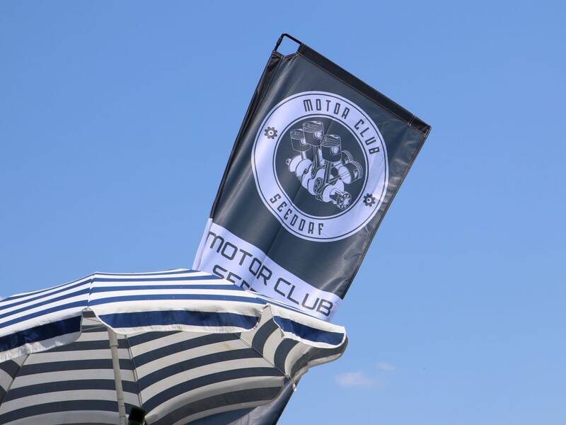 Ein gestreifter Sonnenschirm steht vor einer wehenden Flagge mit dem Emblem des Motor Clubs Seedorf, vor blauem Himmel.