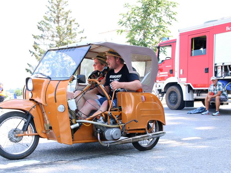 Zwei Personen fahren in einem orangefarbenen, dreirädrigen Fahrzeug mit Verdeck auf einer Straße, im Hintergrund steht ein rotes Feuerwehrauto und ein Mann sitzt am Straßenrand.