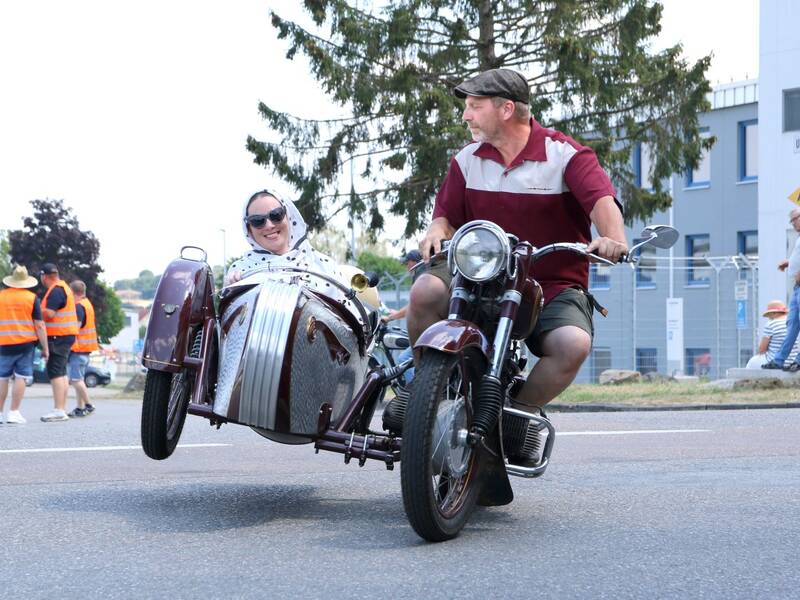Ein Mann fährt auf einem alten Motorrad mit Beiwagen, in dem eine lachende Person sitzt, während im Hintergrund Menschen stehen und zuschauen.