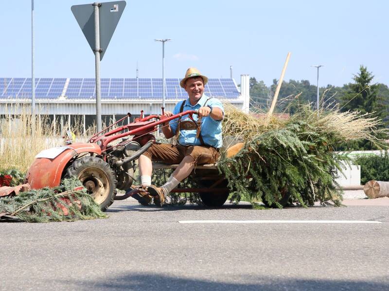 Ein Mann in traditioneller bayerischer Kleidung fährt auf einer ungewöhnlichen Maschine, bestehend aus einem alten Traktor mit Zweigen und Sträuchern, auf einer Straße.