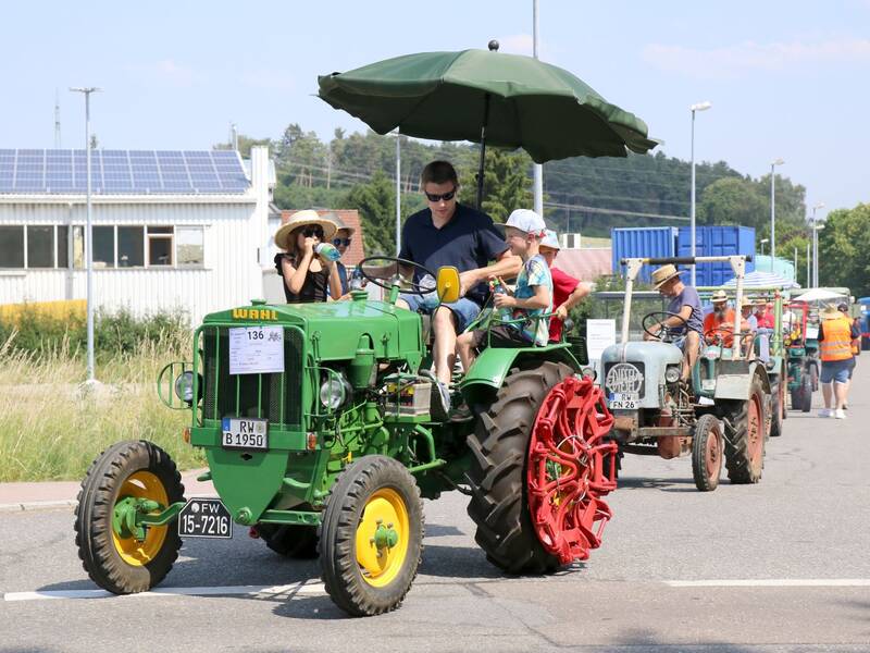 Eine Gruppe von Menschen fährt auf einer Straße in einer Reihe alter Traktoren, angeführt von einem grünen Traktor mit einem Sonnenschirm.
