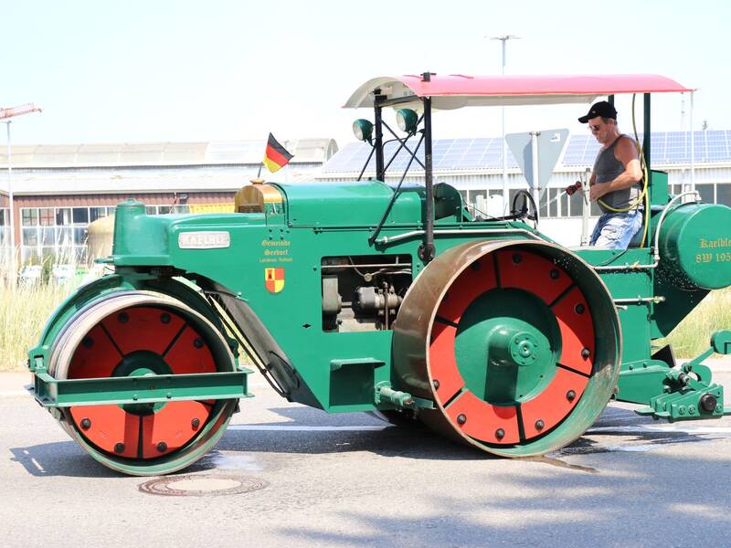 Ein Mann fährt einen grünen historischen Dampfwalze mit rotem Schutzdach auf einer Straße.