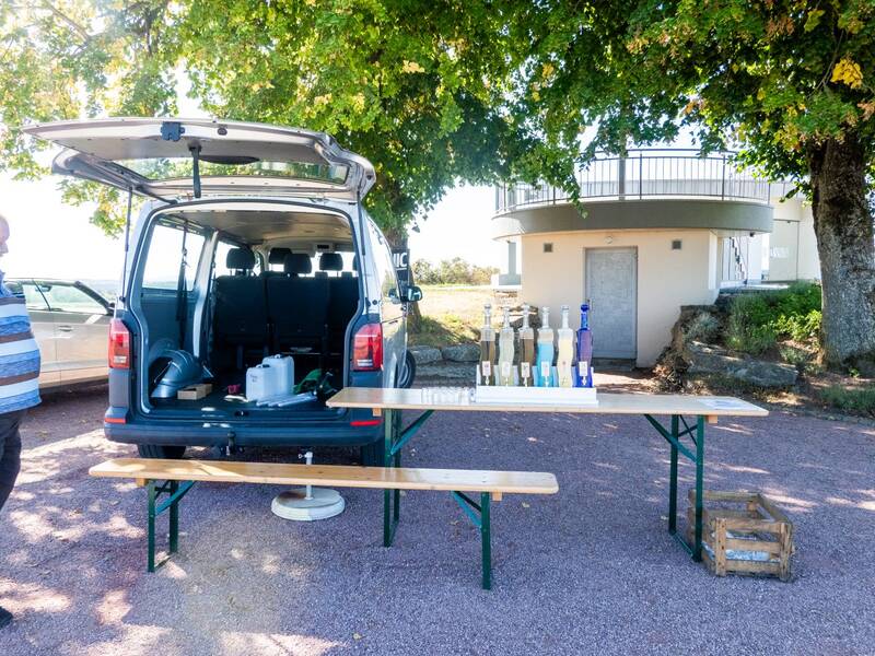 Hinter einem geparkten Auto mit geöffnetem Kofferraum steht ein Tisch mit verschiedenen Flaschen, daneben ein Mann.