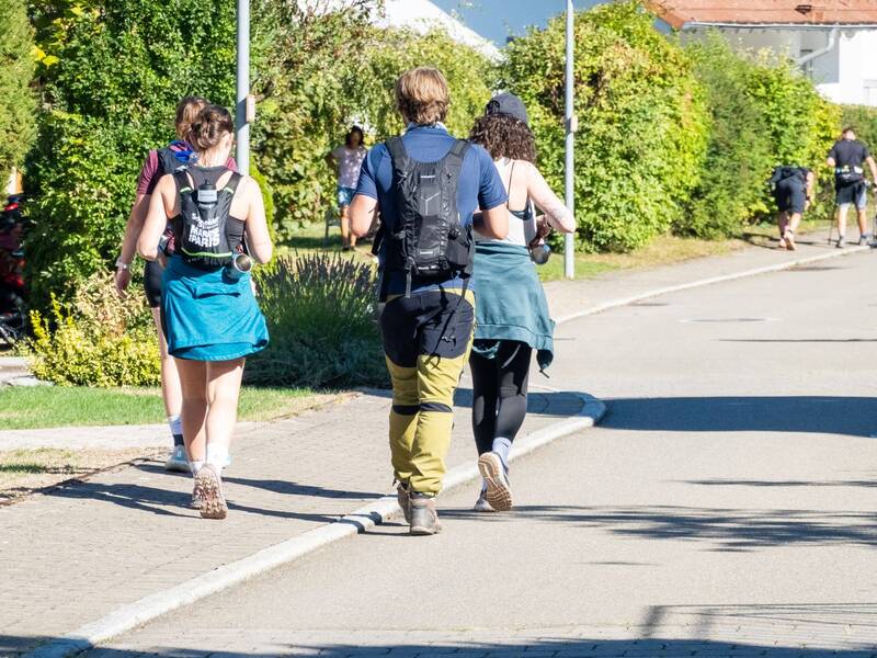 Eine Gruppe von Menschen mit Rucksäcken spaziert auf einem Gehweg entlang einer von Bäumen und Büschen gesäumten Straße.
