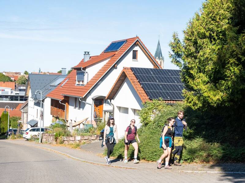 Eine Gruppe von Wanderern geht eine sonnige Straße entlang, vorbei an Häusern mit Solarpanelen auf den Dächern.