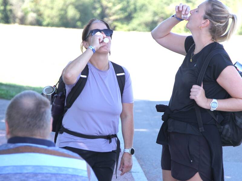 Zwei Frauen mit Rucksäcken trinken während einer Wanderung etwas aus kleinen Bechern, während ein Mann im Vordergrund den Rücken zur Kamera hat.