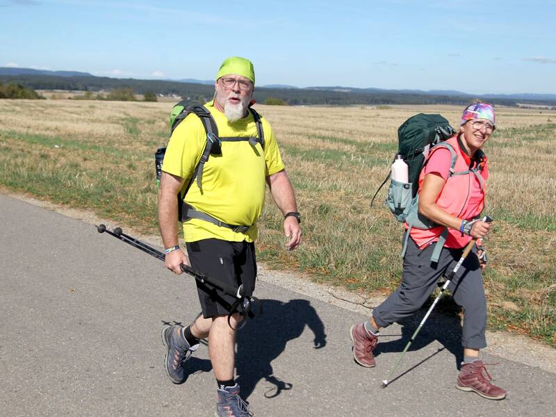 Zwei wandernde Personen mit großen Rucksäcken und Trekkingstöcken gehen auf einem Weg neben einem weiten Feld unter blauem Himmel.