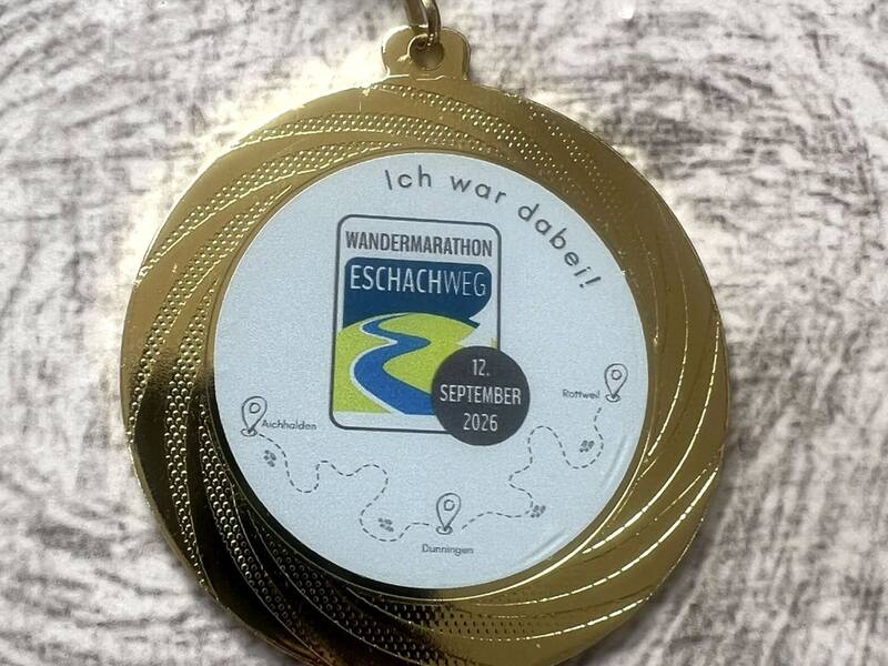 Die Abbildung zeigt eine goldene Medaille mit blauem Band, die den "Wandermarathon Eschachweg" am 12. September 2026 ehrt, und eine Karte mit Stationen wie Aichhalden und Rottweil zeigt.