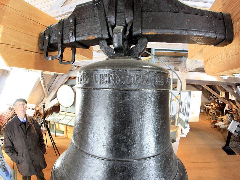 In einem Dachgeschoss mit Holzbalken steht ein Mann neben einer großen, alten Glocke und betrachtet sie.