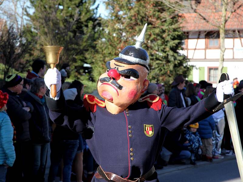 Eine Person in einer Soldaten-Kostümierung mit großer Karnevalsmaske und Helmglocke posiert auf einer Straße, während sich im Hintergrund Menschen versammelt haben.