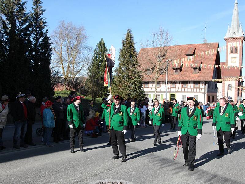 Viele Menschen stehen an einer Straße und schauen einem Umzug zu, bei dem bunte Wimpel über der Straße hängen.