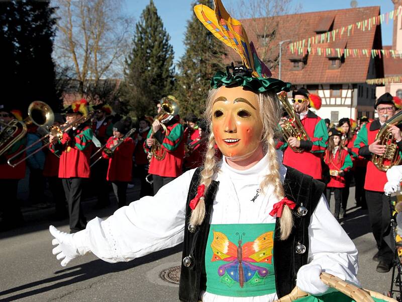 Eine Person in einem bunten Kostüm mit Maske vor eine Musikgruppe in rot-grüner Uniform bei einem Straßenumzug.