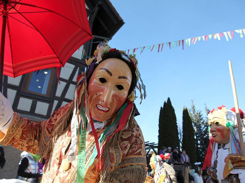 Ein kostümierter Mensch mit einer Holzmaske hält einen roten Regenschirm, während hinter ihm weitere verkleidete Menschen und bunte Girlanden zu sehen sind.
