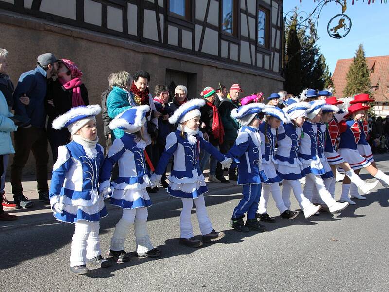 Eine Gruppe von Kindern in blauen Kostümen marschiert während einer Parade die Straße entlang, beobachtet von einer Menschenmenge am Straßenrand.