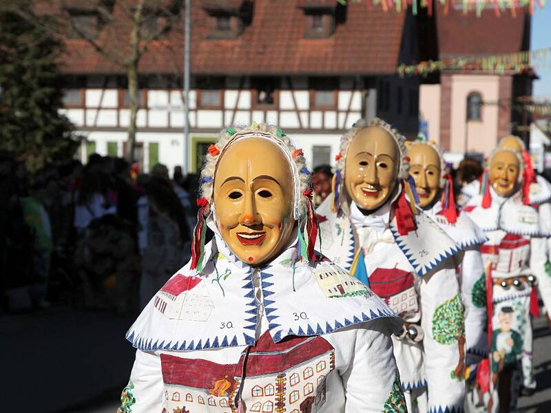Eine Gruppe von Menschen in weißer Kleidung und bunten Masken marschiert während eines Karnevalsumzugs in einer Straße mit traditionellen Fachwerkhäusern im Hintergrund.