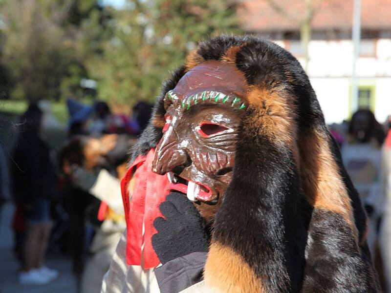 Eine Person trägt eine auffällige Holzmaske mit grimmigem Ausdruck und Pelzumrandung bei einer Veranstaltung im Freien.