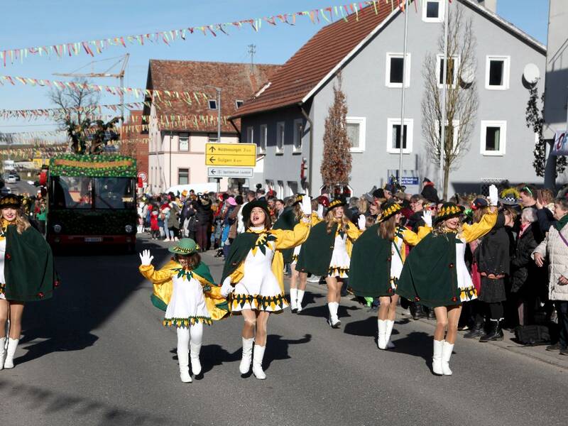 Eine Gruppe von Frauen in weiß gelben Kostümen und traditionellen Hüten marschiert in einer Parade durch eine belebte Straße.