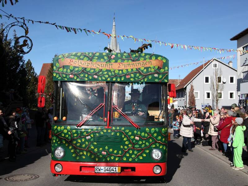 Ein bunt dekorierter Bus fährt durch eine Straße, die von festlich gekleideten Menschen gesäumt ist.
