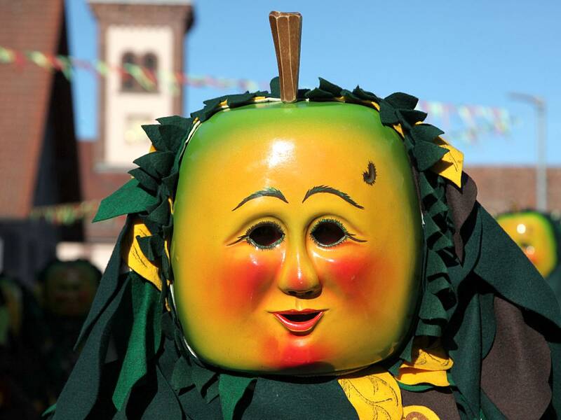 Eine Person trägt eine grüne Maske mit roten Wangen, die einem Apfel ähnlich sieht, umgeben von dunklem, blattartigem Stoff.