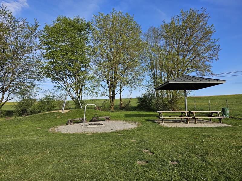 Eine grüne Wiese mit einem Spielplatz, der aus einer Schaukel, einem Sandbereich und einer überdachten Sitzbank besteht, umgeben von Bäumen.