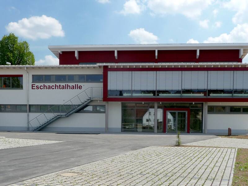 Das Bild zeigt ein modernes Gebäude namens "Eschachtalhalle" mit einer breiten Glasfront, roten Akzenten und einer Außentreppe an der linken Seite.