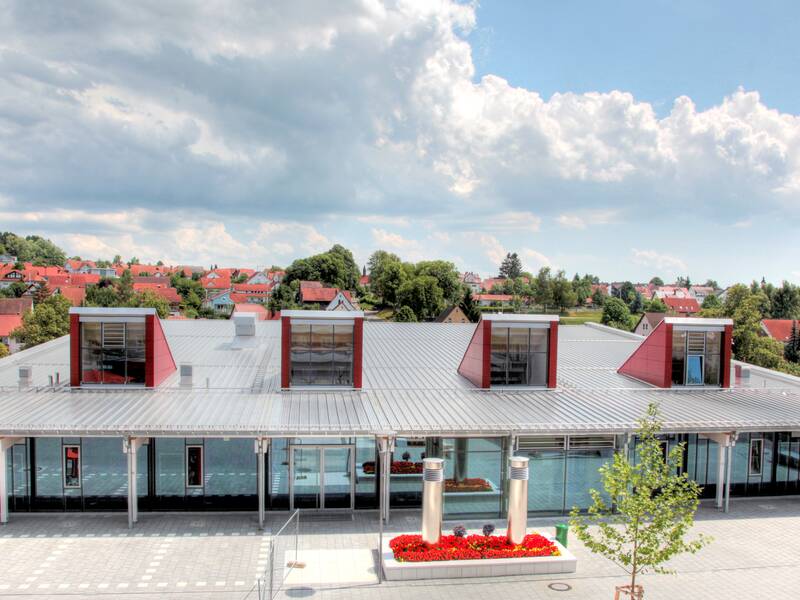 Ein modernes Gebäude mit einem Flachdach und drei rechteckigen Fenstergauben steht vor einer Wohnsiedlung mit roten Ziegeldächern unter einem bewölkten Himmel.