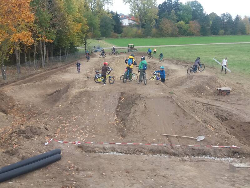 Eine Gruppe von Kindern mit Helmen fährt auf einer hügeligen Fahrradstrecke im Freien, umgeben von herbstlichen Bäumen.
