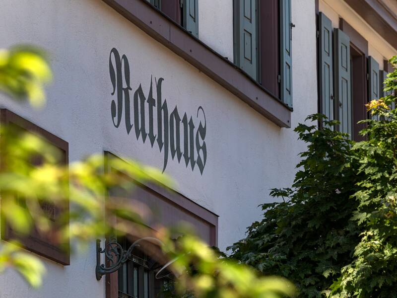 An der Wand eines Gebäudes mit grünen Fensterläden steht in großer Schrift "Rathaus", umgeben von grünen Bäumen.