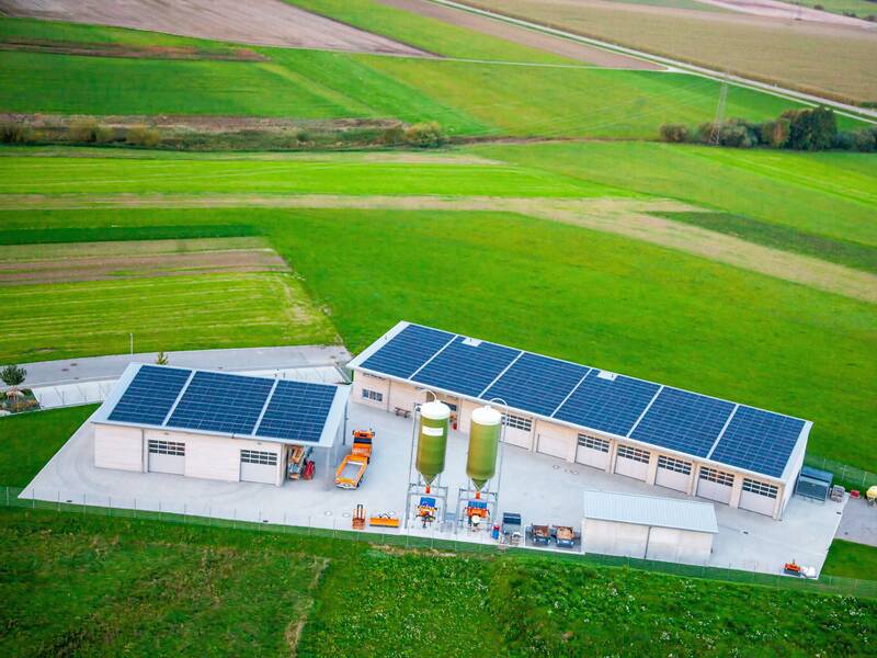 Luftaufnahme eines Gebäudekomplexes mit Solarpaneelen auf dem Dach, umgeben von weiten grünen Feldern.