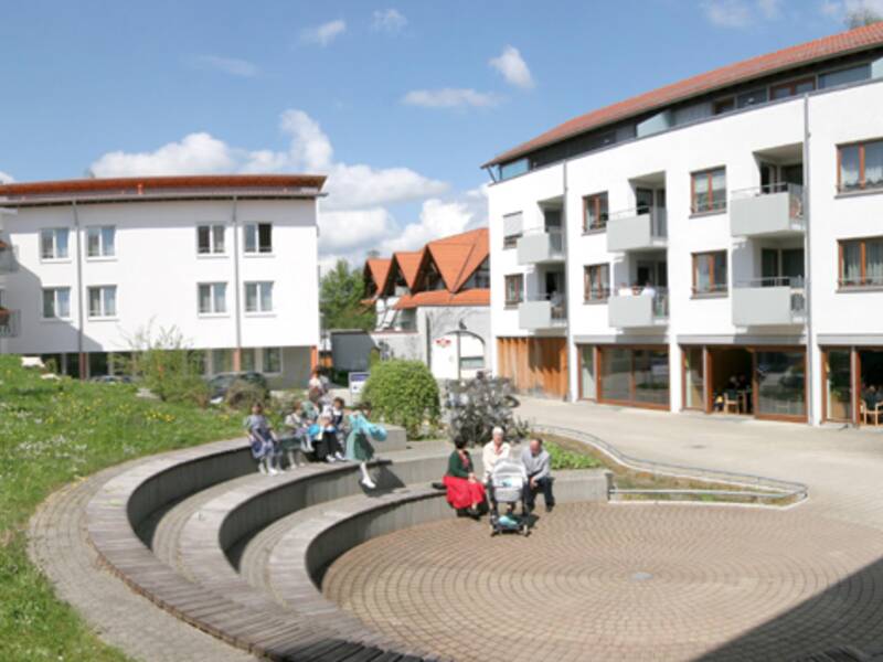 Eine Gruppe von Menschen sitzt und steht auf einem grünen Rasen vor einem modernen Gebäudekomplex mit Treppen und Terrasse unter klarem Himmel.