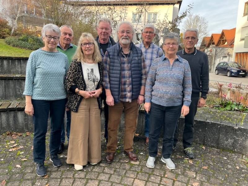 Eine Gruppe von acht älteren Menschen steht auf einem gepflasterten Platz mit einem Gebäude und Vegetation im Hintergrund.