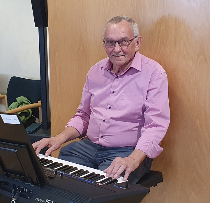 Ein älterer Mann in einem rosa Hemd spielt auf einem Keyboard.
