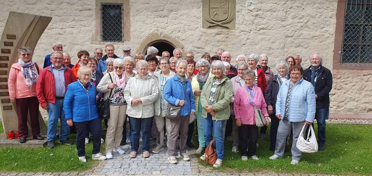 Eine Gruppe älterer Menschen steht dicht beieinander auf einem gepflasterten Weg vor einer historischen Steinmauer.